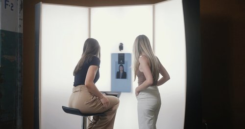 Two women with long hair looking at a screen with a portrait of a woman and a small robot on a bright-lit wall.