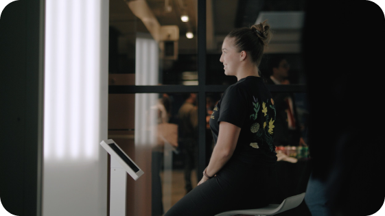 A woman with hair in a bun sitting in front of a touchscreen kiosk in a modern, indoor setting.