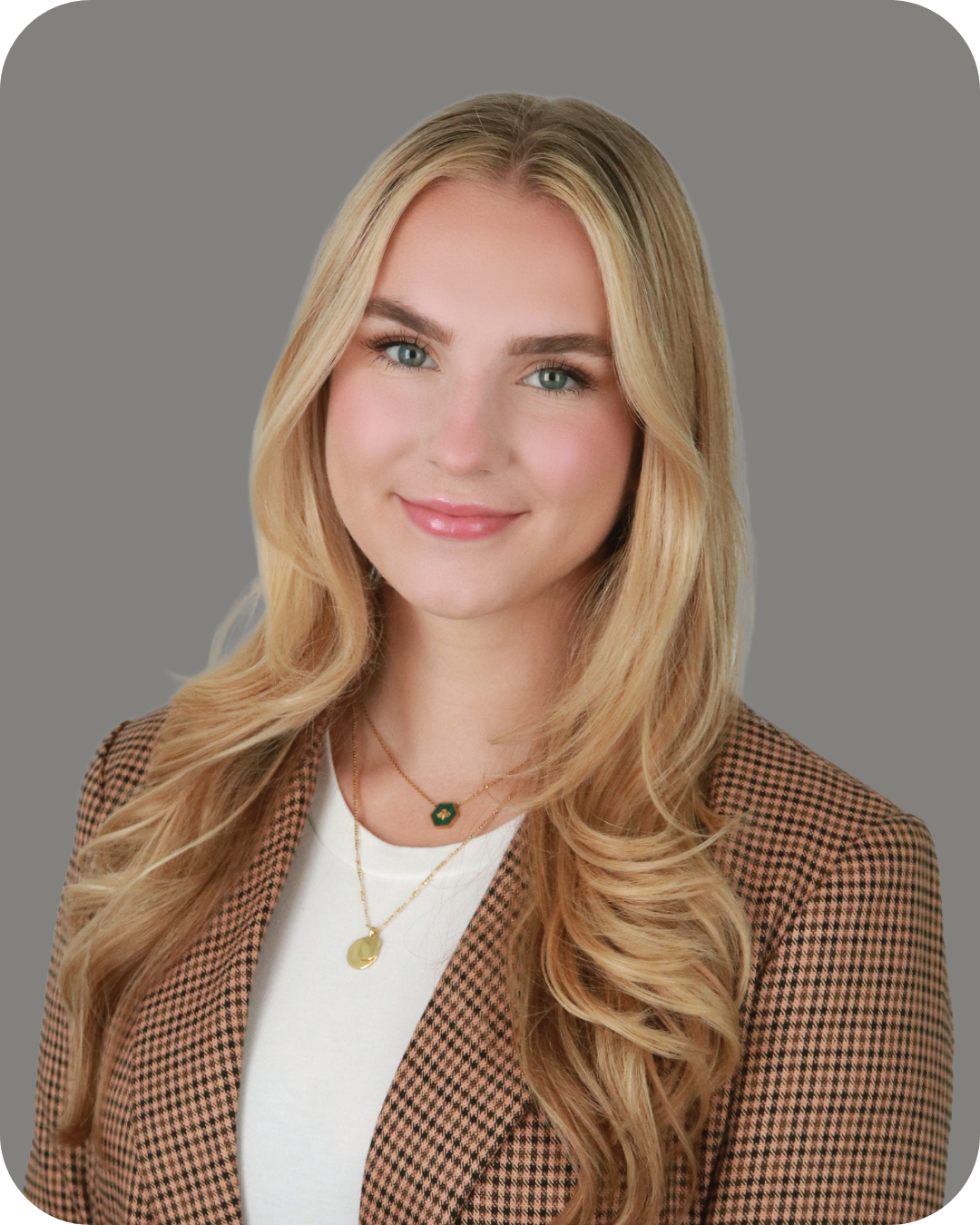 A woman with long, wavy blonde hair, blue eyes, and a light complexion smiling, wearing a checkered blazer over a white top, and layered gold necklaces with pendants, against a neutral gray background.