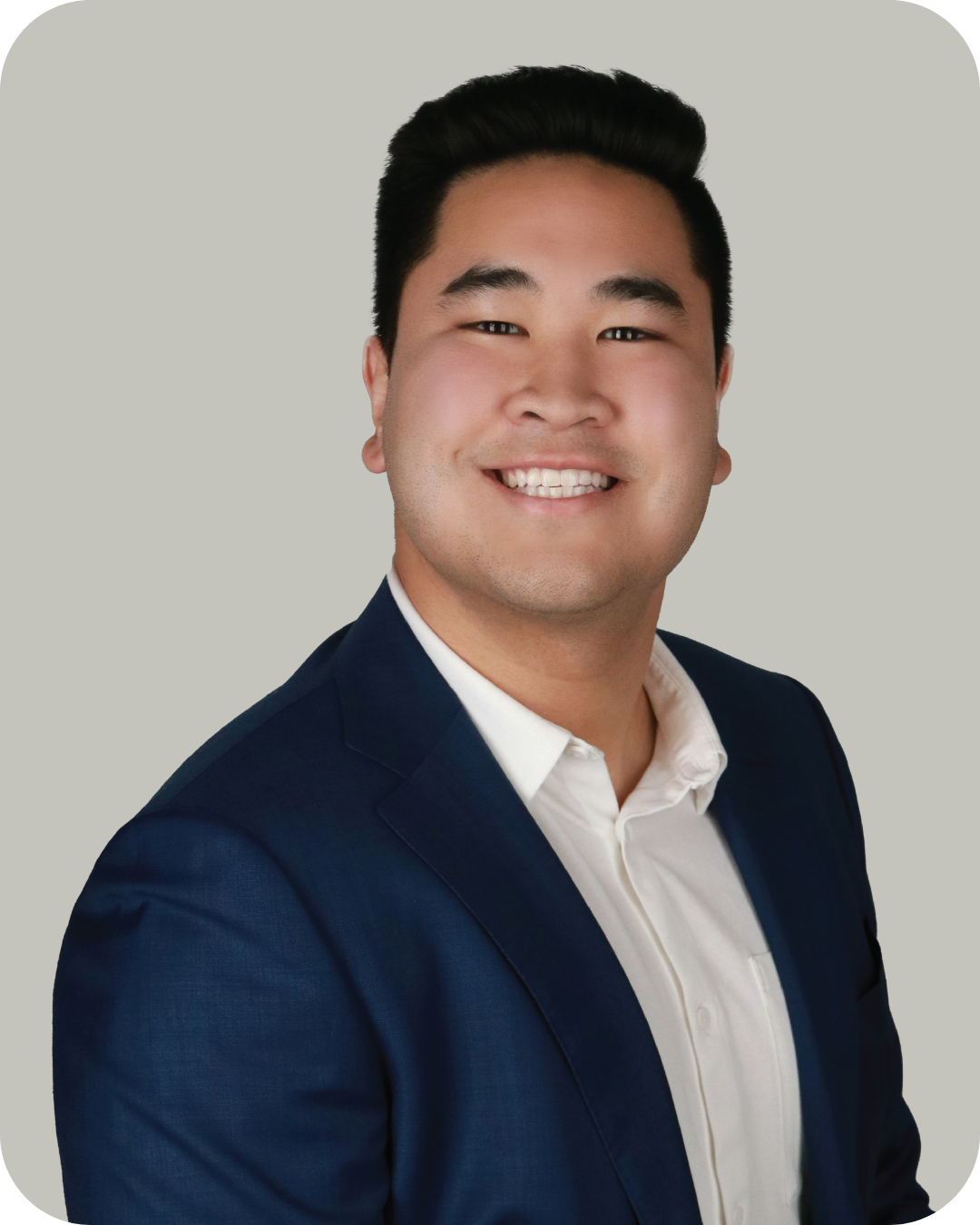 A young man with short black hair and a bright smile, wearing a navy blazer and white shirt, posing against a plain light gray background.