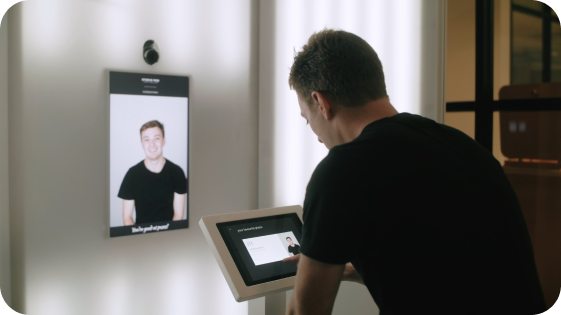 A man interacts with a touchscreen kiosk in front of a facial recognition display showing his photo.