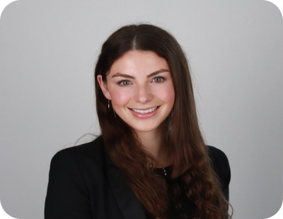 A young woman with long brown hair smiling, wearing a black blazer, against a gray background.