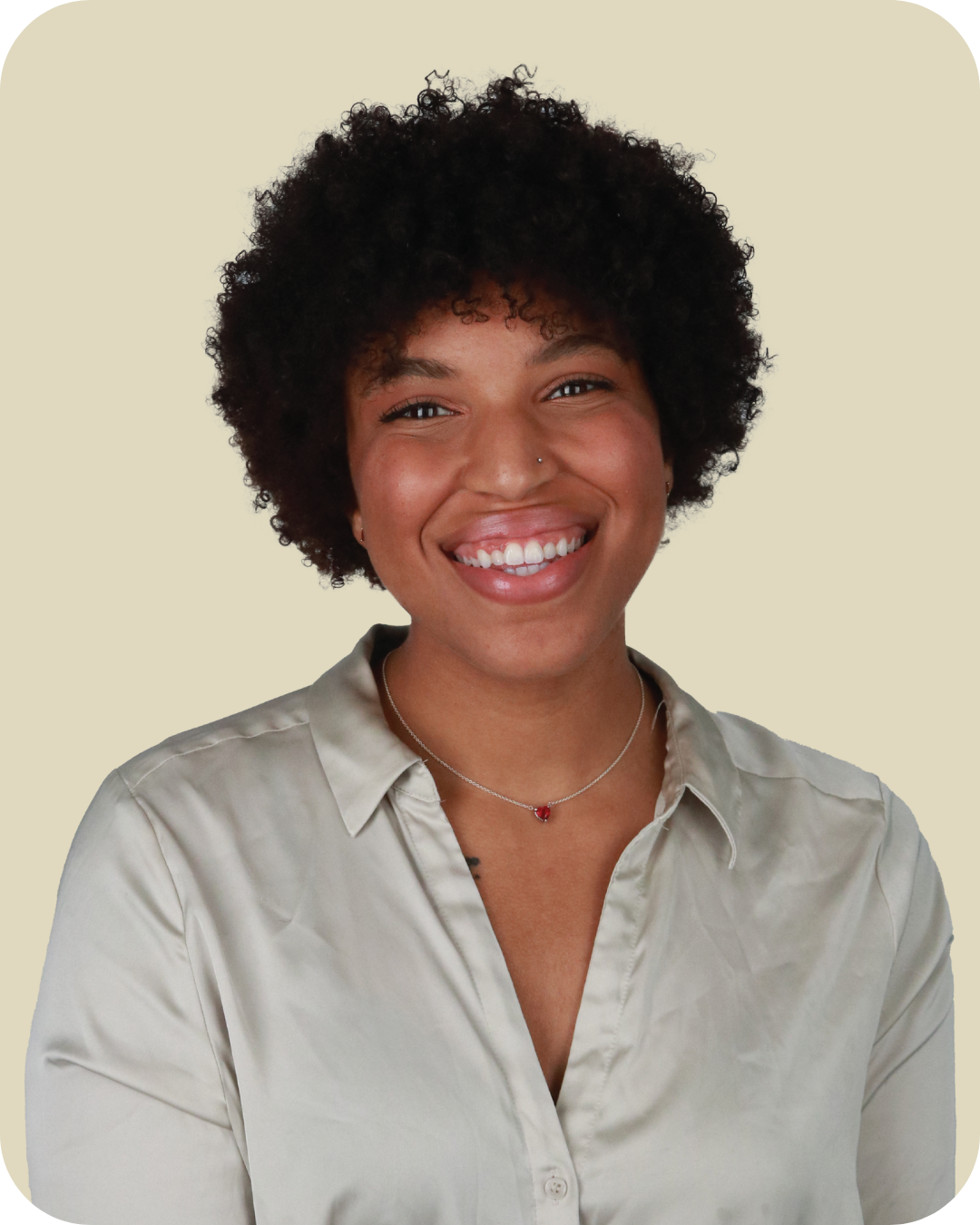 A smiling woman with short curly hair, wearing a light-colored satin blouse and a delicate necklace with a small red pendant, against a plain light-colored background.