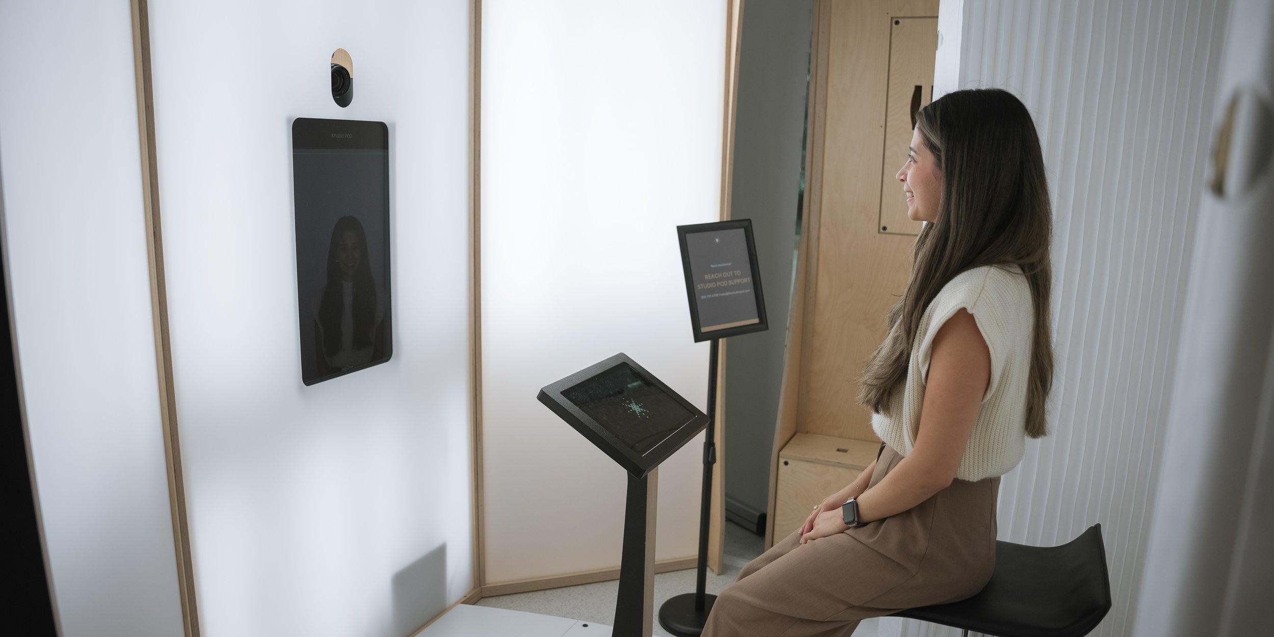 Woman sitting in front of a large screen in a testing or consultation room, with a digital device and informational signs nearby.