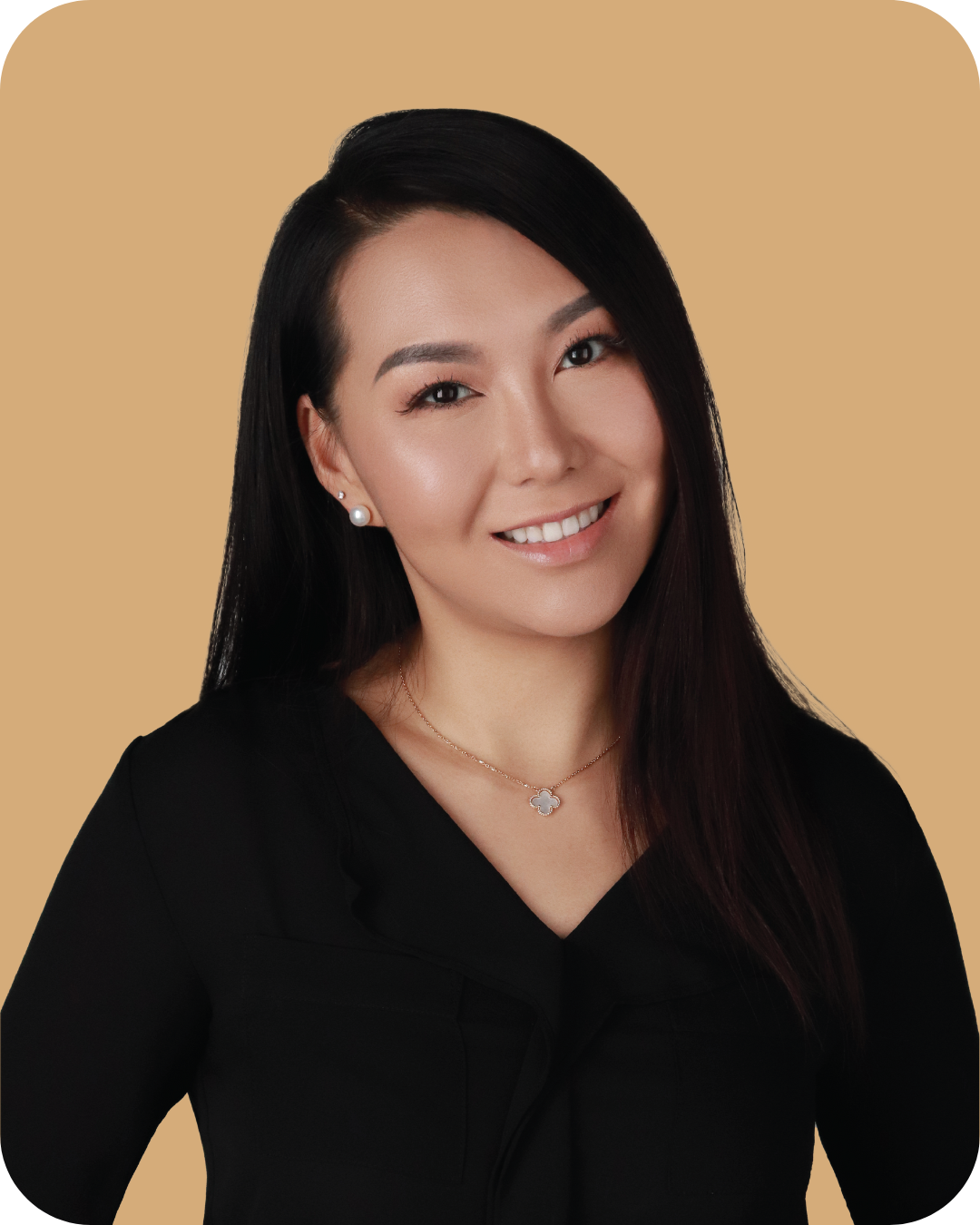 Portrait of a smiling woman with long black hair, wearing a black top, pearl earrings, and a delicate necklace, standing against a beige background.