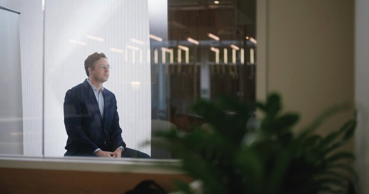 A man with curly hair and glasses in a dark suit and light shirt, seated on a bench in an office, looking thoughtful.