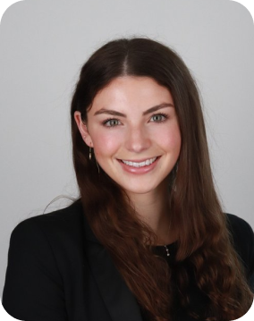 A young woman with long brown hair, smiling, wearing a black blazer, against a plain background.